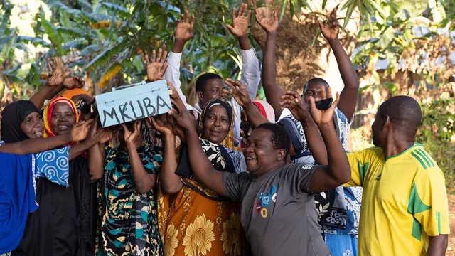 A group of people cheer and celebrate as they hold up a small savings box on which is written 'MKUBA'.