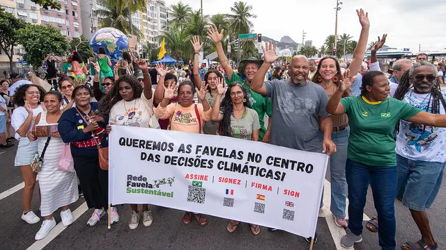 A group of men and women, cheering and chanting and waving their hand in the air, pose behind a sign in Brazilian Portuguese that translates as "We want the favelas to be at the heart of climate decisions".