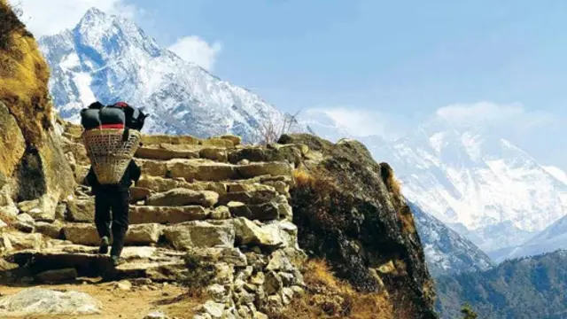 A Nepalese man carrying a large sack on his back walks up some steps with the Himalayas soaring behind. 