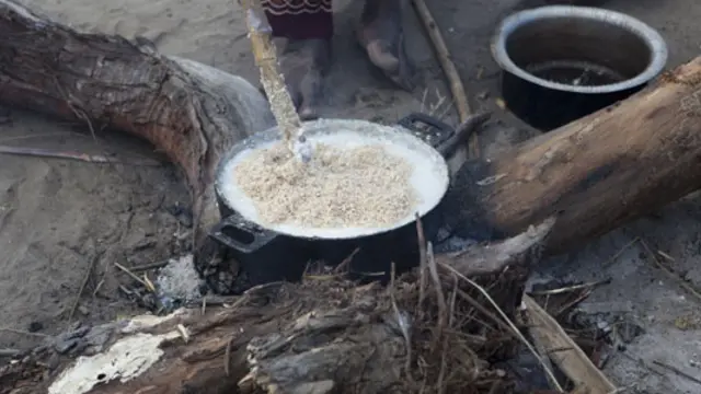 A pot with bubbling food sits cooking on an open fire.