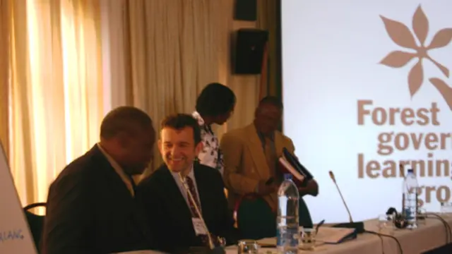 Two men sat behind a desk chat to one another, with the Forest Governance Learning Group logo in the background