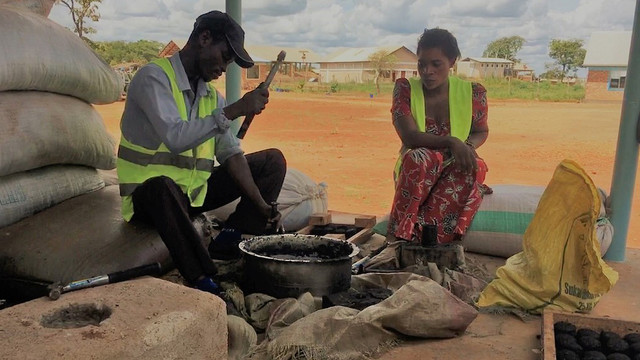 A man and woman sat down cooking with a charcoal cookstove.