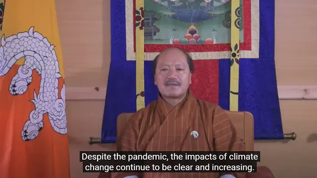 A seated man gives a speech to a camera directly in front of him, next to the Bhutan national flag