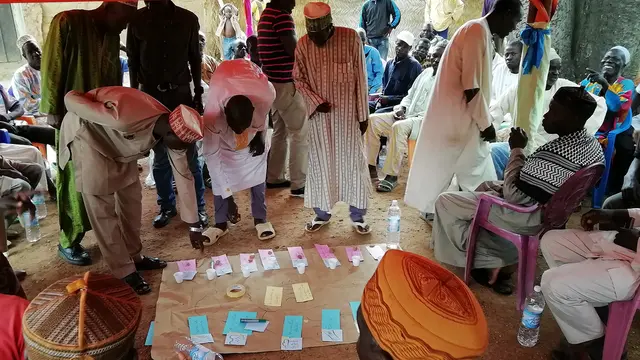 A group of people outside huddle around diagrams and charts laid out on the floor