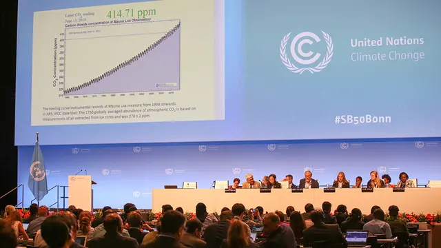 A panel of people sit at a desk facing seated guests, in front of a screen showing a steeply rising graph