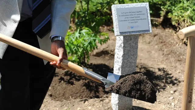 Close-up of a shovel, on which dirt is balanced, during a tree-planting ceremony, with a plaque in the background