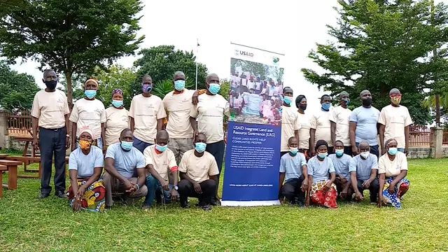 A group of people pose for a photo outside, with trees in the background. There is a banner in the middle