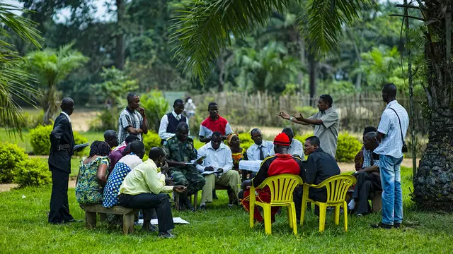 Group of people sit and stand in a circle under a palm tree