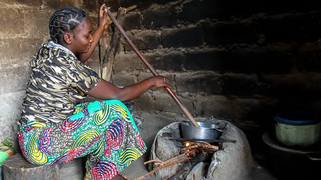 Woman stirs a long stick in a pan on a fireplace made of stone