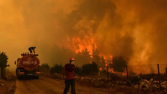 A wildfire burns a forest. A man climbs a fire engine and another one, wearing a mask, walk on the road. 