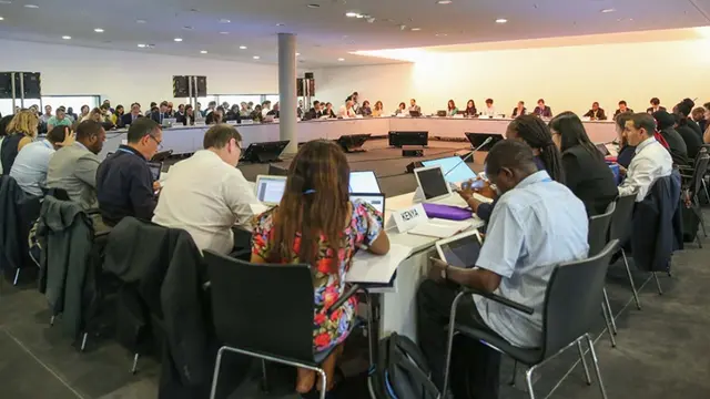 A large number of people, with backs to the camera, are seated around a table. On the tables there are cards indicating which countries they represent