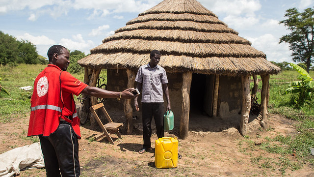 Man stands next to a hut and a jerrycan. Another man, wearing red, points to the right.