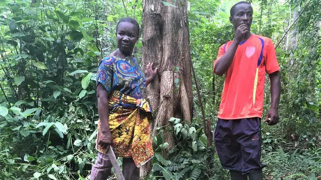 A man and a woman holding a machete stand in front of a tree in a forest