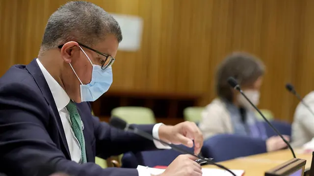 Man wearing a mask is sat at a table with a microphones, and looks at notes.