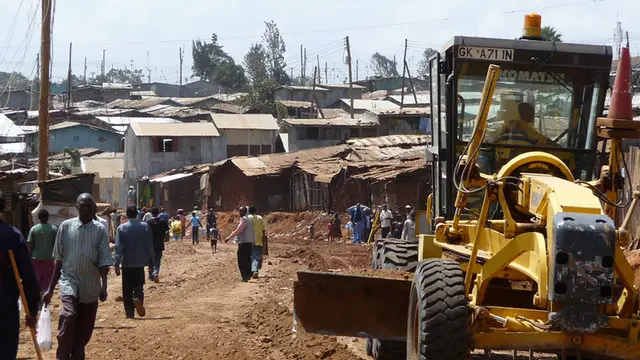 Large yellow digger vehicle parked in front of slum dwellings. 