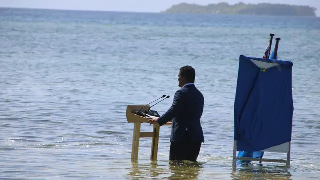 Man stands behind a lectern in the sea to give a speech, with the water above his knees.