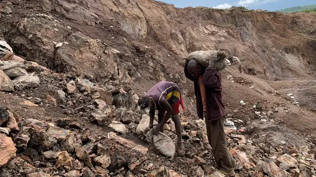 Two men on the side of a mountain; one bends over to pick up two sacks, the other carries them on his shoulders