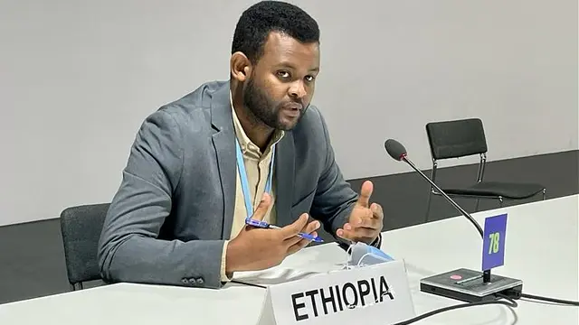 A man sat behind a desk and sign labelled 'Ethopia' gesticulates with his hands