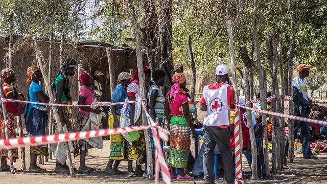 Women stand in a line with empty bags. A man wears a white vest with a red cross on the back.