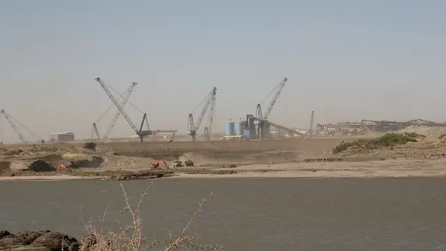 Cranes tower over the construction of a dam, with a body of water in the foreground