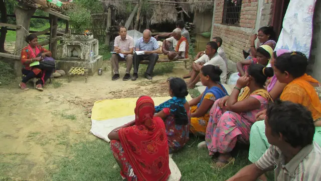 Two men sit on a bench and take notes, while a small crowd sits on the floor in front of them.