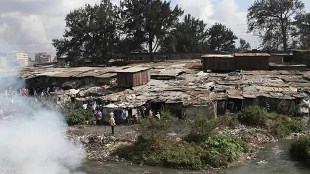 Closely built shacks. In the background, trees. In the foreground, a river and, on the other side of it, smoke.