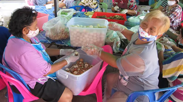 Two women wearing face masks are sitting at a table covered in boxes and bags of vegetables. Together they are preparing food.