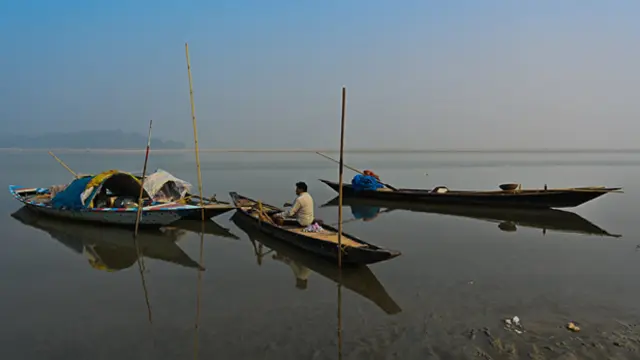 In a broad river, a man sits in a small, narrow boat. Other similar boats are nearby, and poles are sticking up out of the water.