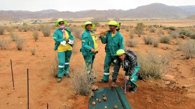 A group of four men and women wearing overalls, one with an axe over his shoulders and one crouching down, pose for a photo. In the foreground are a few rocks, with a dusty landcsape and mountains in the background.