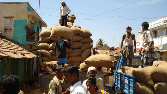 Men stand on lorries loading and unloading sacks at a busy market.