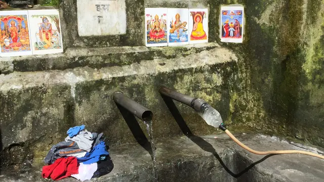Water pours from two pipes set into stone, surrounded by photos, alongside a pile of clothes.