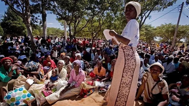 Woman stands in front of a large number of woman and children sat on the floor outside.