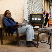 A man sitting in an armchair chair on the street reading a newspaper with a television and a motorbike in the background.