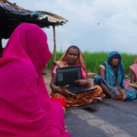 Group of women wearing saris sit on a rug. One of them is holding a laptop.