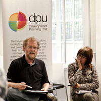 A seated man talks to a group, while a women alongside watches on. Behind them is a board advertising DPU - the Development Planning Unit at UCL.