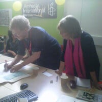 A man leans over a table and writes on a sheet of paper, and a woman watches him.