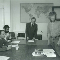 Four men, two standing, two seated, around a table in a boardroom. 