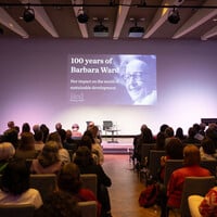 A view form the back of a large hall, over the heads of a seated audience, as a video plays on a big screen.