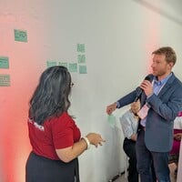 Two people with microphone, standing in front of a room of people, discussing ideas written down on post-its and grouping them.