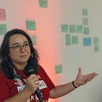 One person with a microphone, standing in front of a room of people and discussing ideas written down on post-its.
