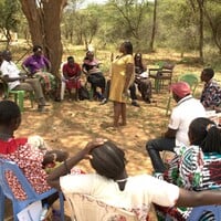 Community members sitting on chairs in a circle, with a person standing in the meeting talking and presenting.