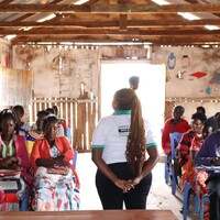 Community members sitting on chairs in a circle, with a person standing in the meeting talking and presenting.