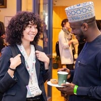 Two people talking in a busy hallway. One person is wearing a white shirt and black blazer and they are smiling. The other person is wearing a traditional dress and hat, holding a cup of coffee.