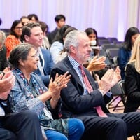 People sitting on chairs, listening to a speech. They are wearing formal clothing.