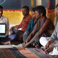A man seated on a rug shows off a laptop screen to other seated men.