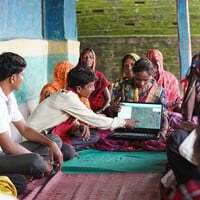 A woman holds up a laptop and a man points to the screen, as people seated all around look interested. 