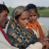 A woman holds up a phone as a man points to something on the screen, while two other women watch on. Water is in the background. 