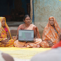 A woman holds up a laptop while talking to a group of others sat outside on blankets.